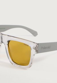 Gafas de sol con montura transparente y clara, lentes amarillos y brazos grises, con la marca "Polaroid" en el lente y en la patilla.