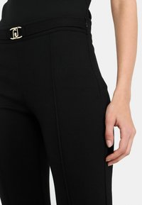 Crne krojene hlače s glatkom teksturom, zlatnim uređajem kopče za remen i bočnim zatvaračem. Elegantan, ravno krojen dizajn.