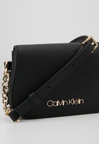 Sac bandoulière Calvin Klein noir avec détail de chaîne dorée et sangle réglable sur un fond neutre.