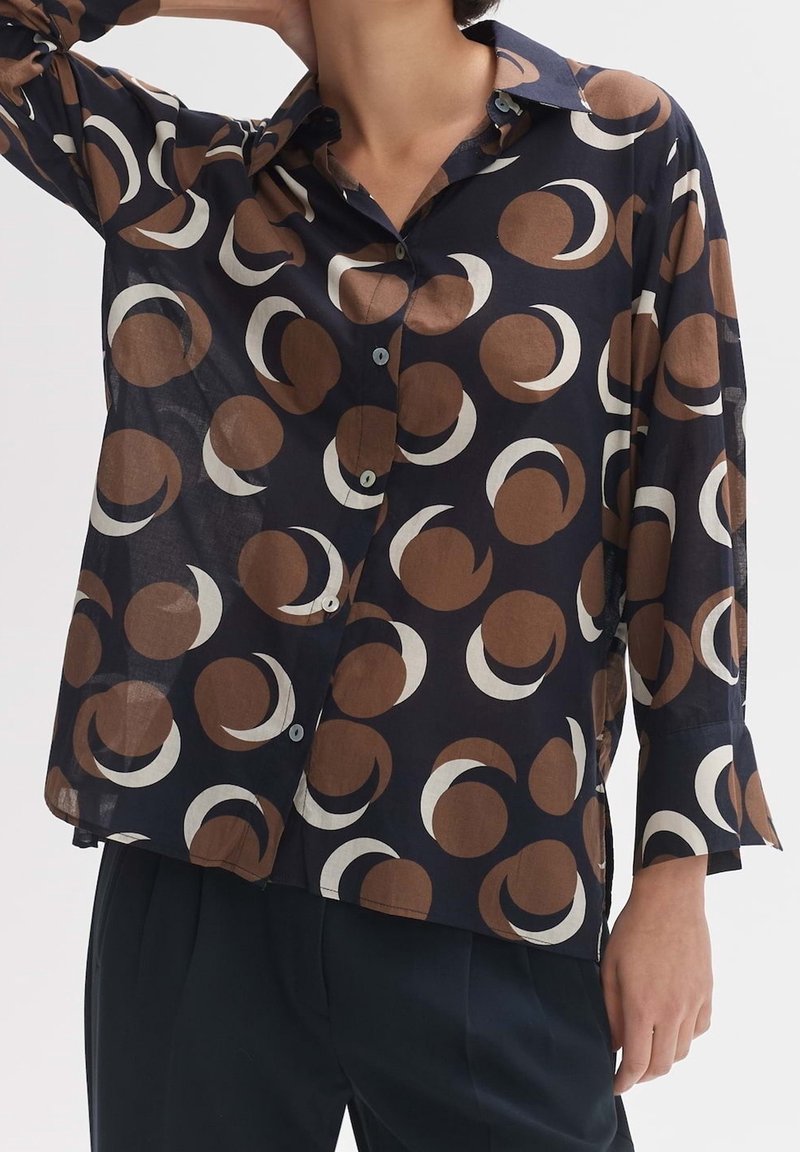 Personne portant une chemise noire à boutons avec un motif circulaire abstrait marron et blanc et un pantalon foncé, un bras levé derrière la tête.
