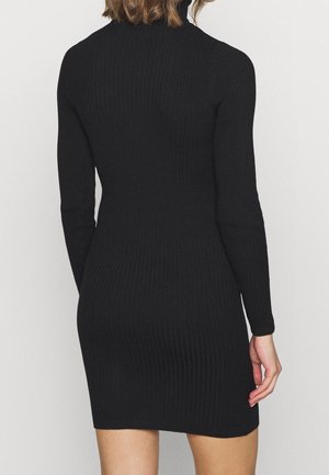 Vrouw die een aansluitende, lange mouwen, zwarte geribbelde gebreide jurk draagt, gezien van achter tegen een effen lichte achtergrond.