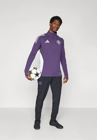 Haut d'entraînement Adidas à manches longues violet avec des accents blancs et gris, associé à un pantalon d'entraînement noir et des chaussures de sport noires, tenant un ballon de football.
