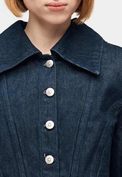Γυναίκα που φοράει σκούρο μπλε τζιν μπουφάν με μεγάλο γιακά και τέσσερα μεταλλικά κουμπιά στο μπροστινό μέρος, κομμένο κάτω από τη μύτη.