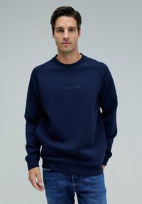 Sudadera azul marino con acentos acanalados en los hombros y logo bordado "SALSA JEANS" en el pecho. Combinada con jeans de mezclilla.