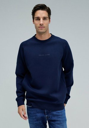 Sudadera azul marino con acentos acanalados en los hombros y logo bordado "SALSA JEANS" en el pecho. Combinada con jeans de mezclilla.