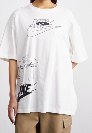 Personne portant un t-shirt Nike blanc oversize avec des logos et du texte imprimés en noir, associé à un pantalon cargo beige.