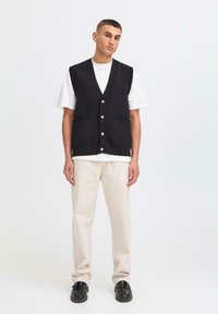 Jonge man in een zwart knoopvest over een wit T-shirt, beige broek en zwarte schoenen, staand tegen een effen witte achtergrond.
