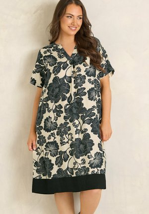 Kvinde iført en knælængde, beige kjole med stort sort blomstermønster og korte ærmer, stående foran en ensfarvet lys væg.