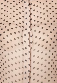 Blusa translúcida con una base de un beige claro y lunares oscuros. Presenta un cierre de botones en el centro y una textura suave y lisa.