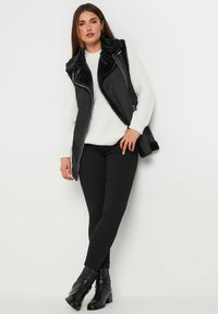 Svart imitationsläderväst med en pälskrage, lager på lager över en vit stickad tröja, matchad med svarta skinny jeans och ankelboots. Slät yta.
