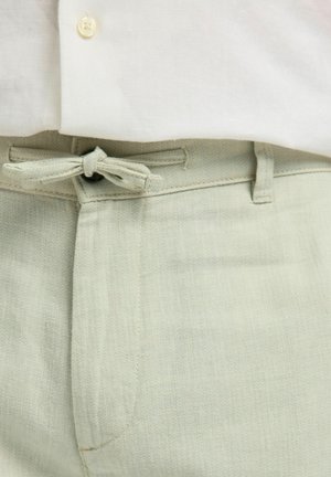 Světlé zelené kalhoty s látkovým páskem uvázaným mašli, nošené s bílou košilí, zobrazující detail pasu a knoflíků.