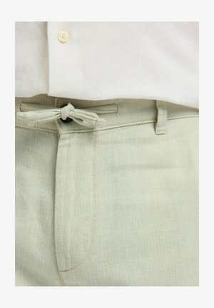 Světlé zelené kalhoty s látkovým páskem uvázaným mašli, nošené s bílou košilí, zobrazující detail pasu a knoflíků.