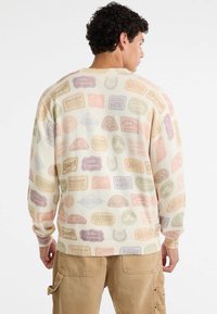 Crème trui met een repetitief grafisch ontwerp featuring verschillende kleurrijke labels. Gebreide textuur met geribde manchetten en zoom.