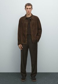 Ung mand står op ad en grå væg iført en mørkebrun nubuckjakke, matchende mørkebrune bukser, en strikket sweater og mørke sko.