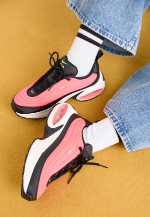 Osoba nosząca różowo-czarne buty sportowe Nike Air z białymi skarpetkami i niebieskimi dżinsami na podłodze w kolorze musztardowym.