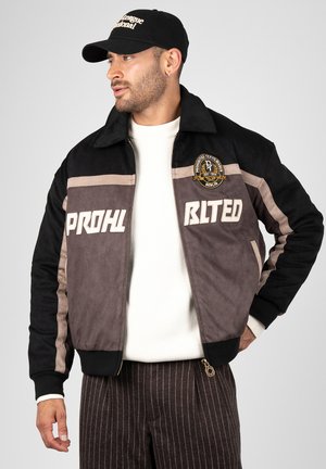 Mand iført en sort og grå jakke med lapper og hvide bogstaver, en hvid sweater, stribede brune bukser og en sort kasket, der kigger mod venstre.