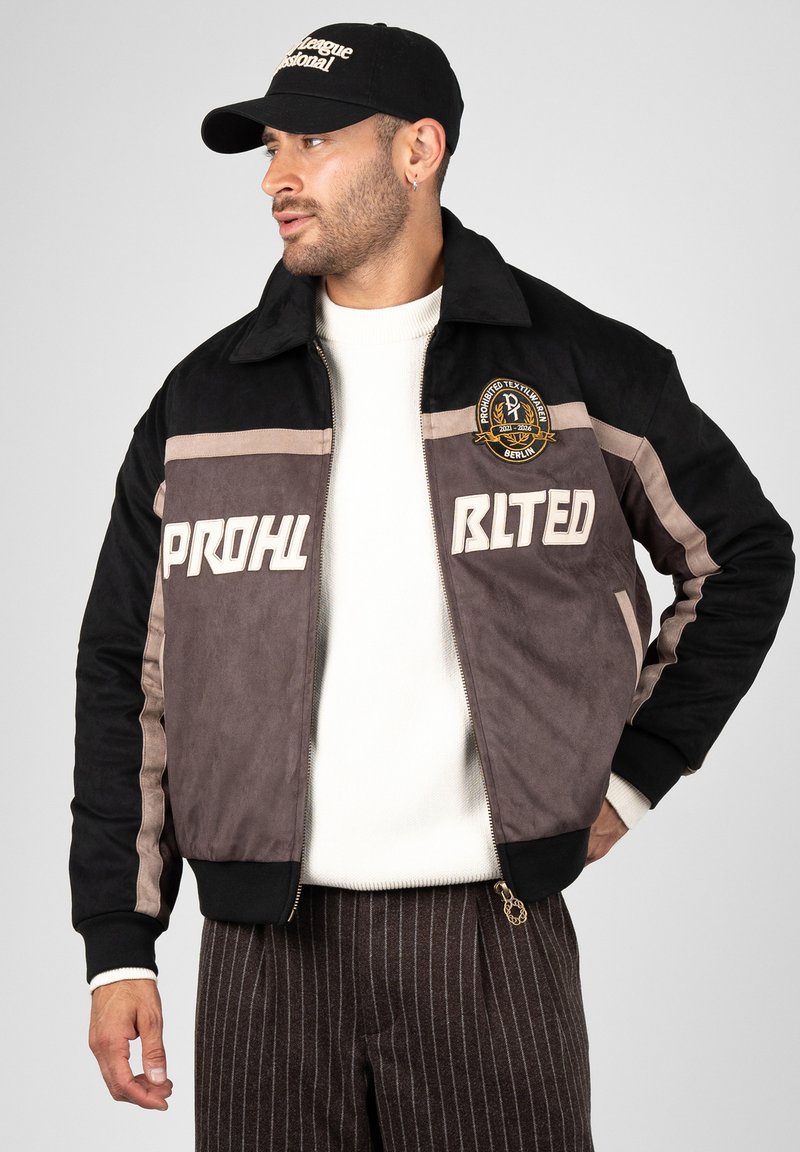 Homme portant une veste noire et grise avec des écussons et des inscriptions blanches, un pull blanc, un pantalon marron rayé et une casquette noire regardant vers la gauche.