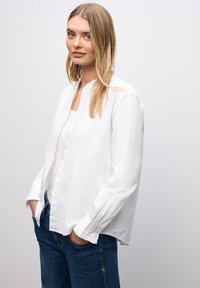 Camisa blanca de botón con cuello redondo y puños con volantes, combinada con jeans azules. La tela parece suave con un diseño de ajuste relajado.