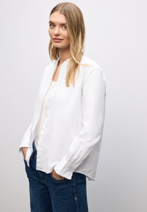 Witte blouse met ronde kraag en ruches bij de mouwen, gecombineerd met blauwe jeans. De stof lijkt glad met een ontspannen pasvorm.
