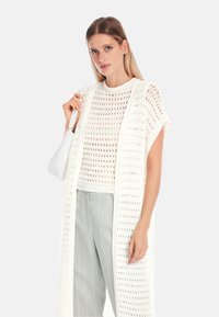 Weißer offen gestrickter Vest über einem kurzen weißen Strickoberteil, kombiniert mit hellgrauen gestreiften Hosen. Die Tasche ist glatt und weiß.