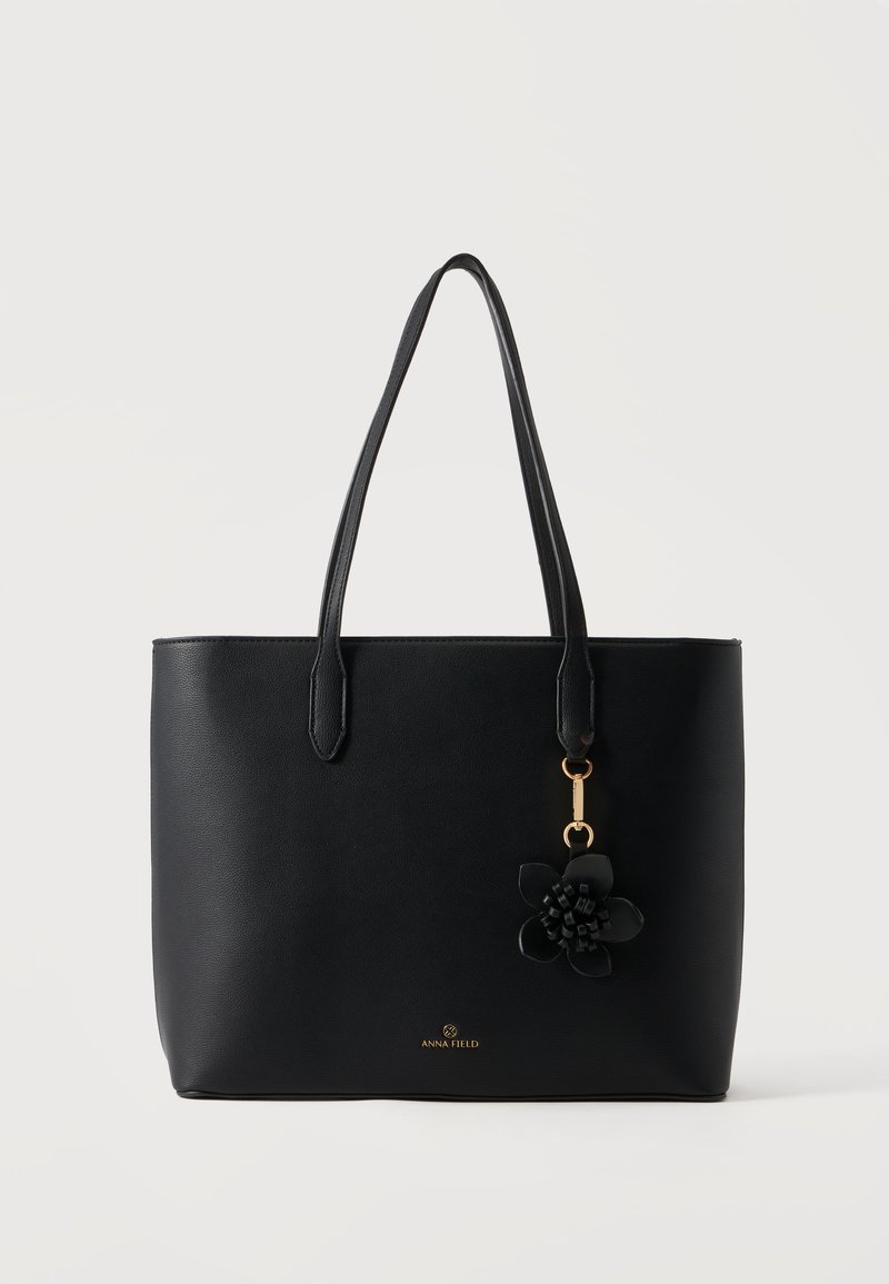 Sac cabas en cuir noir avec deux poignées, petit pendentif fleur et logo doré "Anna Field" sur le devant.