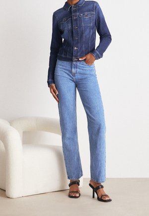 Tmavě modrá džínová bunda s kapsami a knoflíky, kombinovaná s světle modrými džíny s rovnými nohavicemi a černými sandály na podpatku. Jednoduché pozadí.