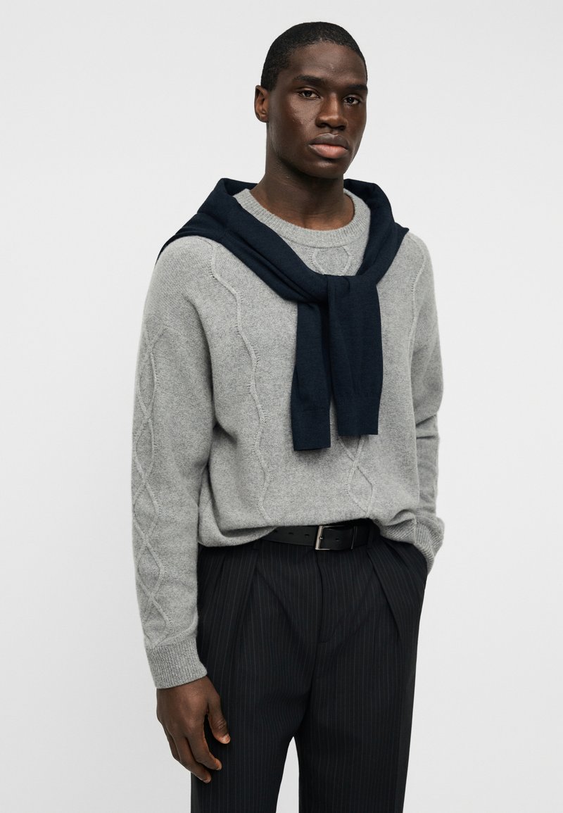Gray knitted sweater with a cable pattern, worn over the shoulders is a dark navy scarf. Black striped trousers, and black belt complete the look.