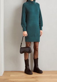 Robe tricotée teal avec col roulé côtelé, manches texturées, accompagnée d'un sac noir et de bottes brunes épaisses, sur un fond neutre.