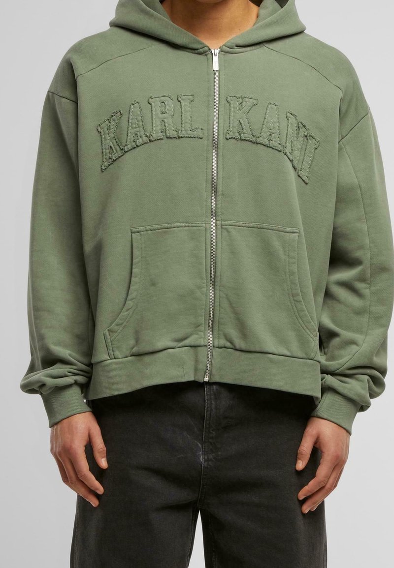 Personne portant un sweat à capuche vert zippé avec le texte "KARL KANI" sur la poitrine et un pantalon noir, sur un fond gris uni.