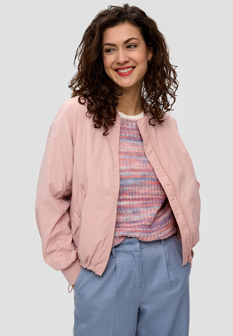 Veste bomber rose clair avec des poignets élastiques, une fermeture éclair et des poches latérales, portée par-dessus un pull en tricot multicolore et un pantalon bleu clair.