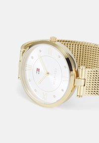 Orologio da polso dorato con quadrante bianco, numeri romani, indicatori in cristallo, bracciale a maglia e piccolo logo Tommy Hilfiger sul quadrante.