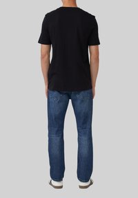 Persoon in een effen zwart T-shirt met korte mouwen, blauwe jeans en witte sneakers, staand met de rug naar de camera op een grijze achtergrond.