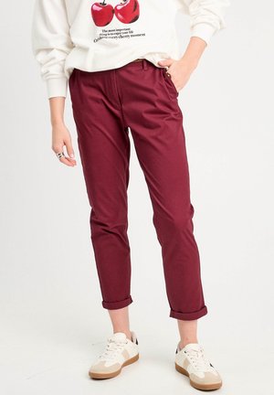 Persoon die opgerolde bordeauxrode broek, witte sneakers en een witte trui draagt met kersgrafieken en tekst.