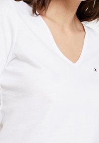 Camiseta blanca de algodón con escote en V, que presenta un pequeño logo bordado en rojo y azul cerca del cuello. Textura suave, diseño sencillo.