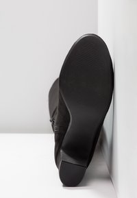Botte en daim noir vue de la semelle, avec une semelle extérieure en caoutchouc lisse et un talon texturé, reposant contre un mur blanc sur un sol blanc.