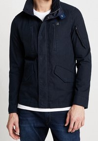 Marinblå jacka med ståkrage, framdragkedja och flera fickor. Tillverkad av ett texturerat tyg, har en liten logotyp på bröstet.