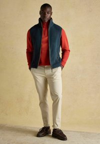 Vīriešu apģērbs, kas sastāv no tumši zilas vestes virs sarkanas rāvējslēdzēja virsjakas, kombinēts ar krēmkrāsas biksēm un tumši brūnām kurpēm. Gludas tekstūras, ikdienišķs dizains.