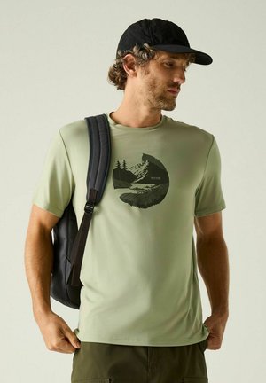 Homme portant un t-shirt vert clair avec un motif montagne, une casquette noire et un sac à dos noir sur une épaule, regardant vers sa droite.