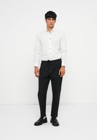 Witte knoopshirt met zwarte knopen, gecombineerd met zwarte joggingsbroek en zwarte schoenen. Platte kraag en slim fit.