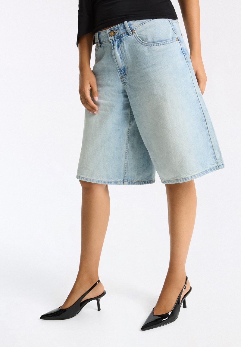 Personne portant un short en jean bleu clair au genou, un haut noir à manches longues et des chaussures slingback à talon kitten en cuir verni noir.