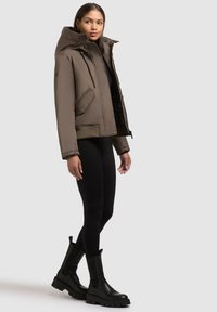 Braune gepolsterte Jacke mit Kapuze, die einen Reißverschluss, Seitentaschen und elastische Bündchen hat, getragen zu schwarzen Leggings und klobigen schwarzen Stiefeln.
