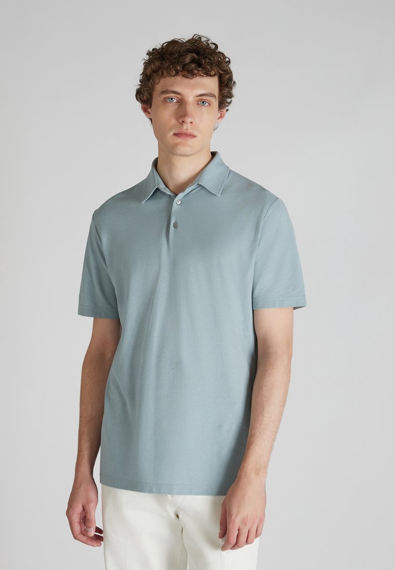 Polo à manches courtes en bleu clair, avec un col, une patte de boutonnage à deux boutons et une texture lisse. Associé à un pantalon écru.