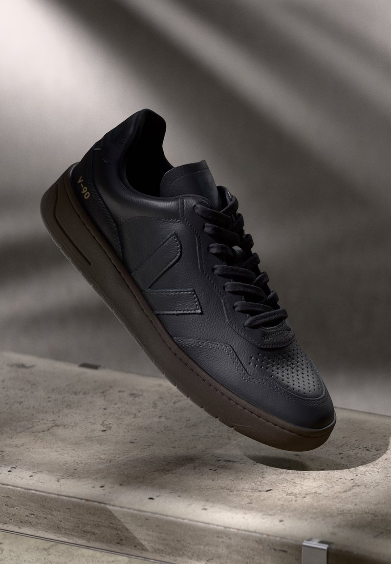 Baskets en cuir noir avec embout perforé, semelle épaisse, lacets et détail du logo, positionnées sur une surface en béton avec un éclairage doux d’en haut.
