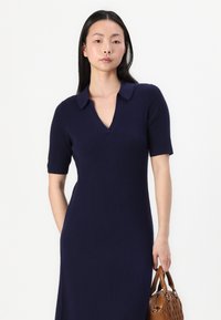 Navy blue, ribbed knit dress with short sleeves and a V-neck collar. Model holds a brown handbag with a textured pattern.