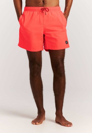 Homme debout, pieds nus, portant un short de bain corail vif avec des poches latérales et un logo circulaire noir sur l'ourlet.