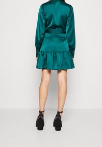 Tyrkysové saténové šaty s dlouhými rukávy, páskem v pase a rozšířeným lemem. Temné podpatky s páskem kolem kotníku a robustním blokovým podpatkem doplňují vzhled.