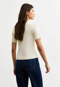 Witte geribde top met korte mouwen en een fitted ontwerp, met een hoge halslijn en een gladde textuur, gecombineerd met donkerblauwe wijde jeans.
