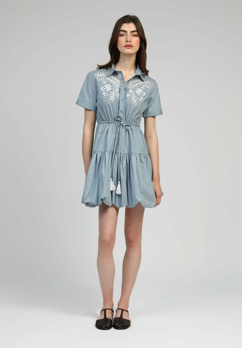 Robe rayée bleu clair avec manches courtes, ourlet froncé, taille avec cordon de serrage, et broderie florale blanche. Comprend des pompons à la taille.