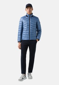 Light blue puffer jacket with a high collar, zip closure, and diagonal quilting. Paired with navy pants and gray sneakers.