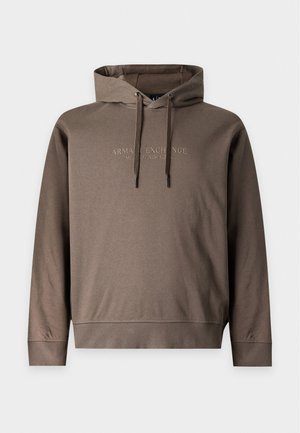 Joven con sudadera marrón con capucha, vaqueros anchos de color azul claro y zapatillas negras, de pie frente a un fondo blanco liso.